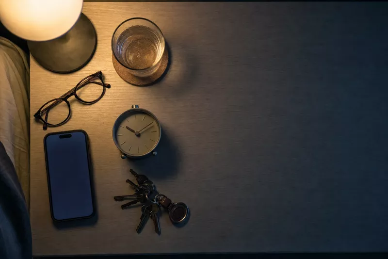 Nachttisch von oben bei Nacht: Handy mit leuchtendem Bildschirm, Glas Wasser, Brille, Schlüsselbund, Wecker, Lampe